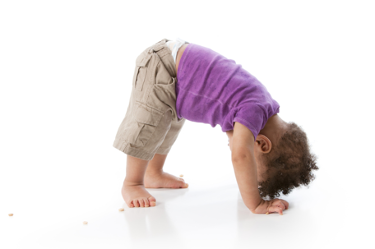 A full length image of a Black or African American toddler boy playing and tumbling. He is bending over, looking upside down, ready to do a somersault.